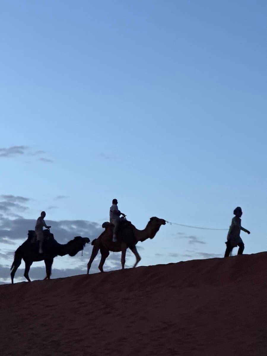 A guide leads two riders on camels along a dune ridge at dusk