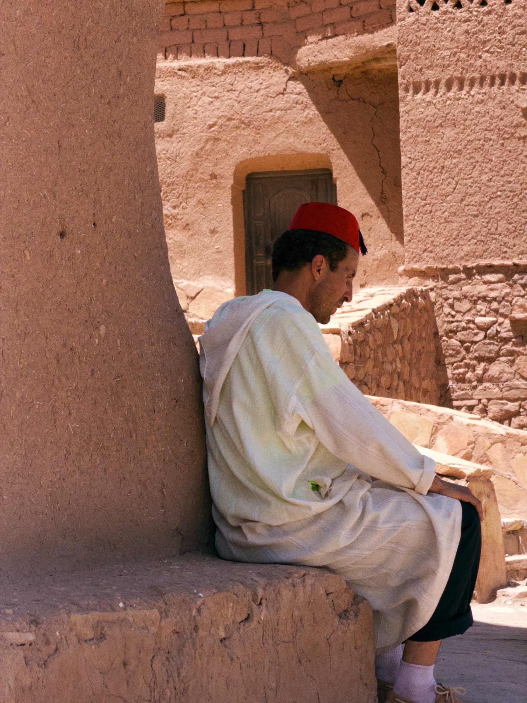 A man in a white djellaba and red fez resting by a kasbah wall